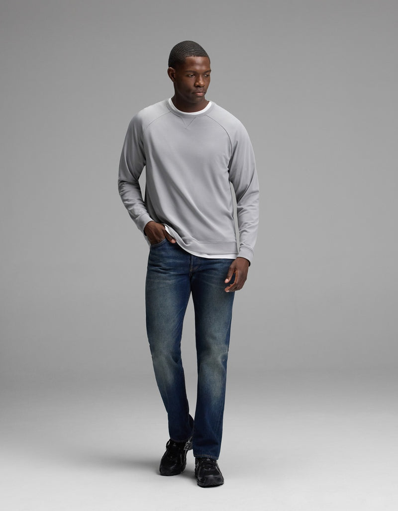 A man wearing the Cozy Earth Men’s Brushed Bamboo Pullover Crew in light gray, paired with blue jeans and black shoes, stands against a plain light gray background, looking to his right with one hand in his pocket.