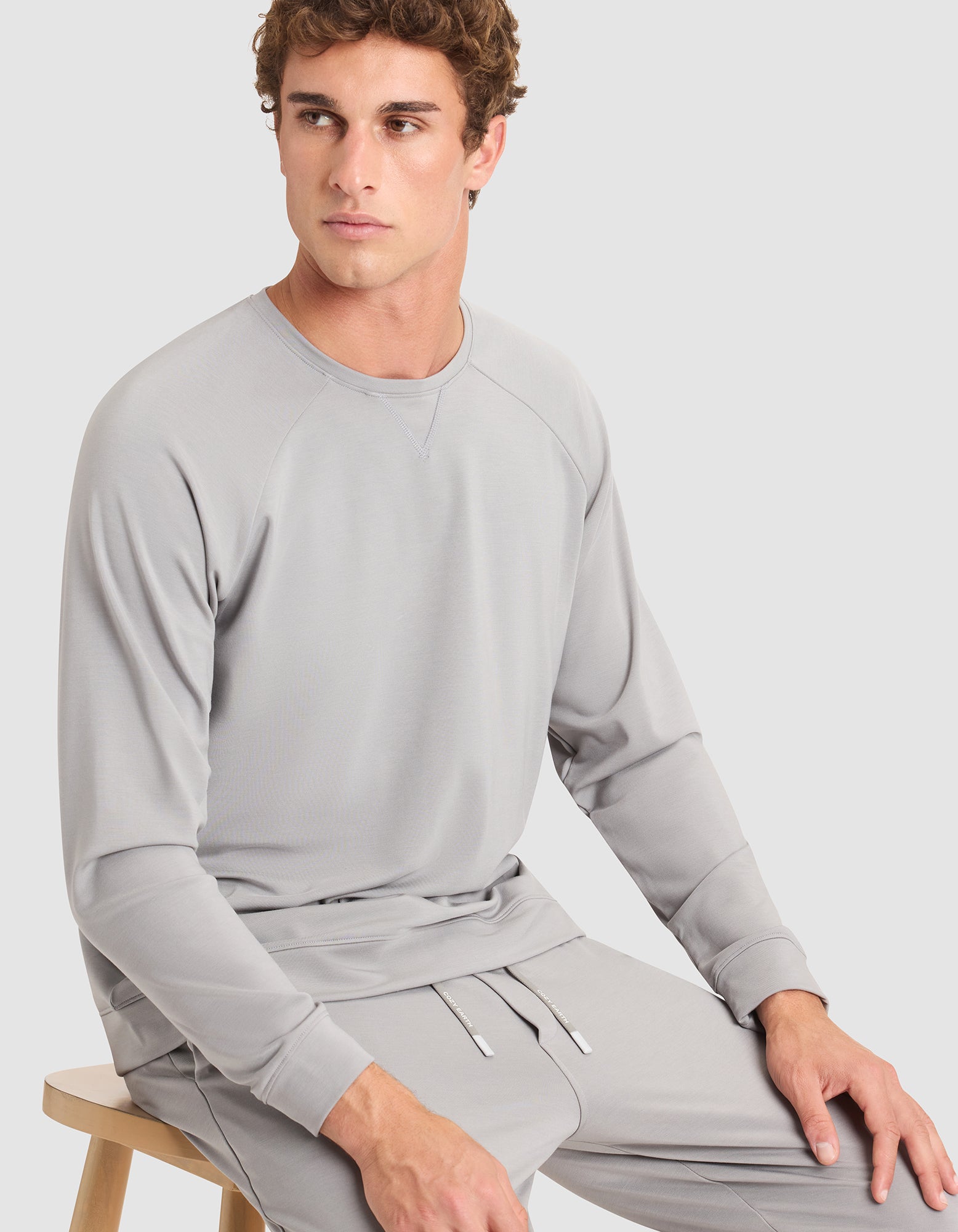 A young man with curly hair, dressed in a light gray Cozy Earth Men's Brushed Bamboo Pullover Crew and matching pants, sits on a wooden stool against a plain light background, gazing slightly to his left. 