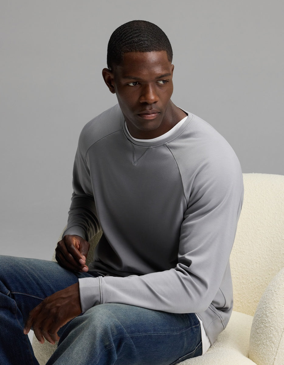Wearing the Cozy Earth Men’s Brushed Bamboo Pullover Crew, a man sits on a cream-colored chair in jeans, gazing to his left against a neutral gray background.