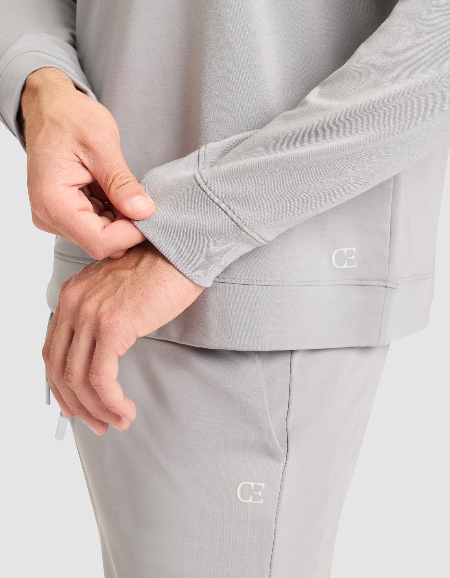A person wearing Cozy Earth’s Men’s Brushed Bamboo Pullover Crew in light gray adjusts the sleeve. The sweatshirt features a small "CE" logo. Only their torso and hands are visible against a plain background. 