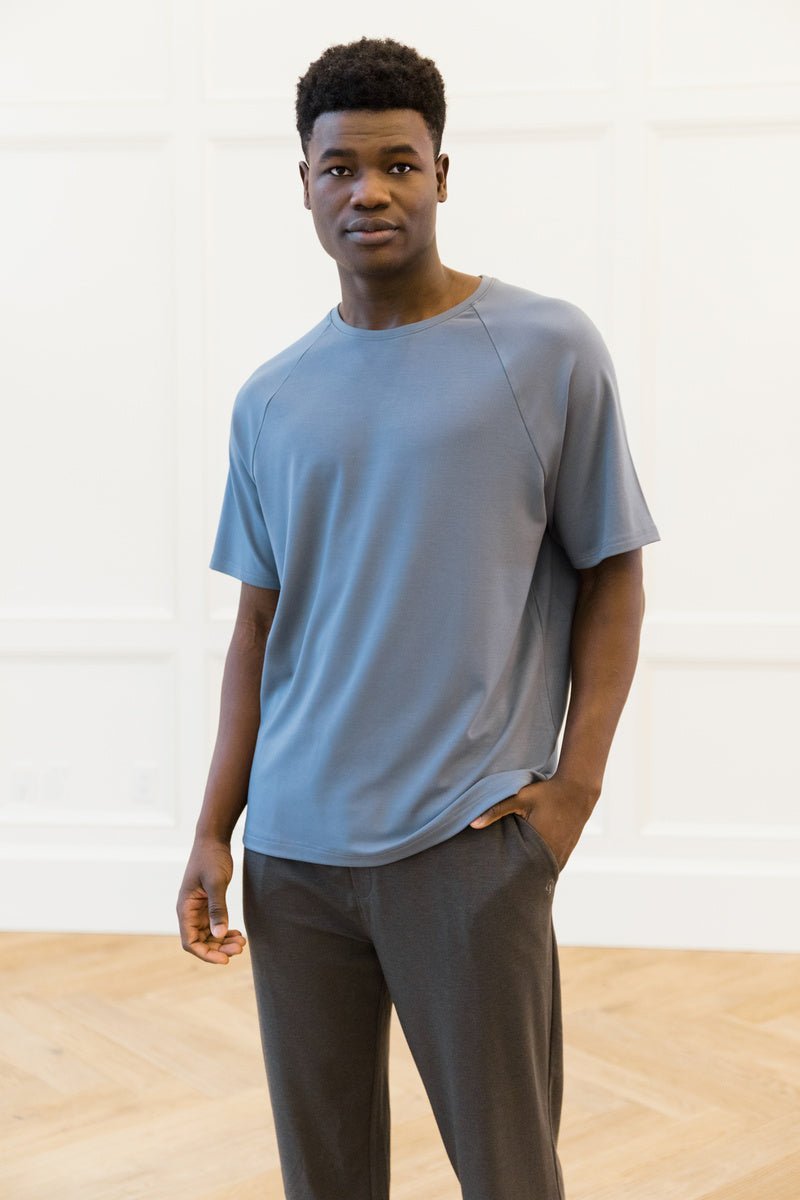 A person stands indoors, hand in pocket, wearing a Cozy Earth Men's Brushed Bamboo Raglan Tee in light blue and dark pants against a light-colored wall over a wooden floor. 