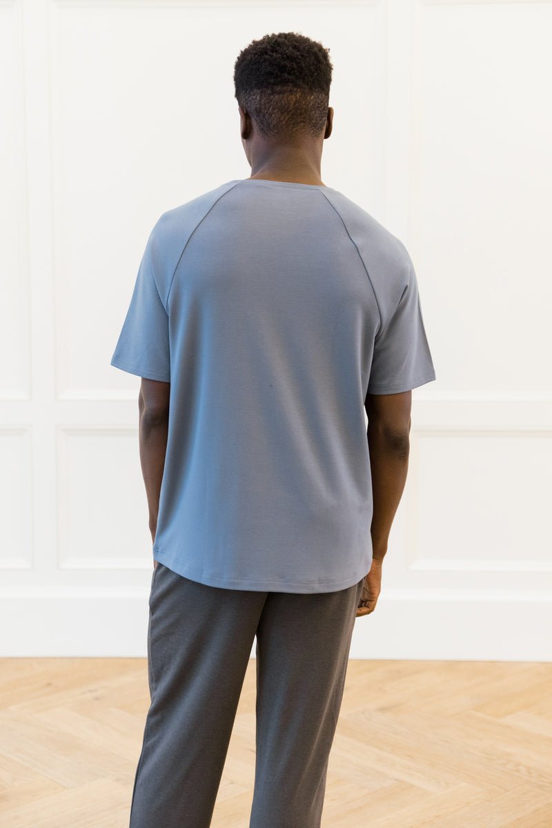 A person with short curly hair, facing away from the camera, is wearing Cozy Earth's Men's Brushed Bamboo Raglan Tee in blue and dark gray pants. The setting includes a white paneled wall and a wooden floor.  