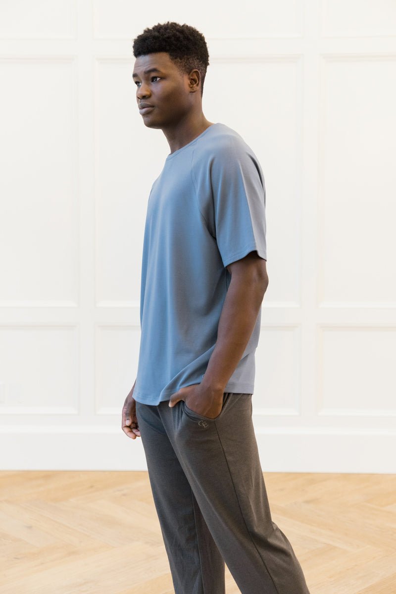 A person in a Cozy Earth Men's Brushed Bamboo Raglan Tee, light blue in color, and dark gray pants stands indoors on a wooden floor with a white paneled wall behind them. They are gazing to their left. 