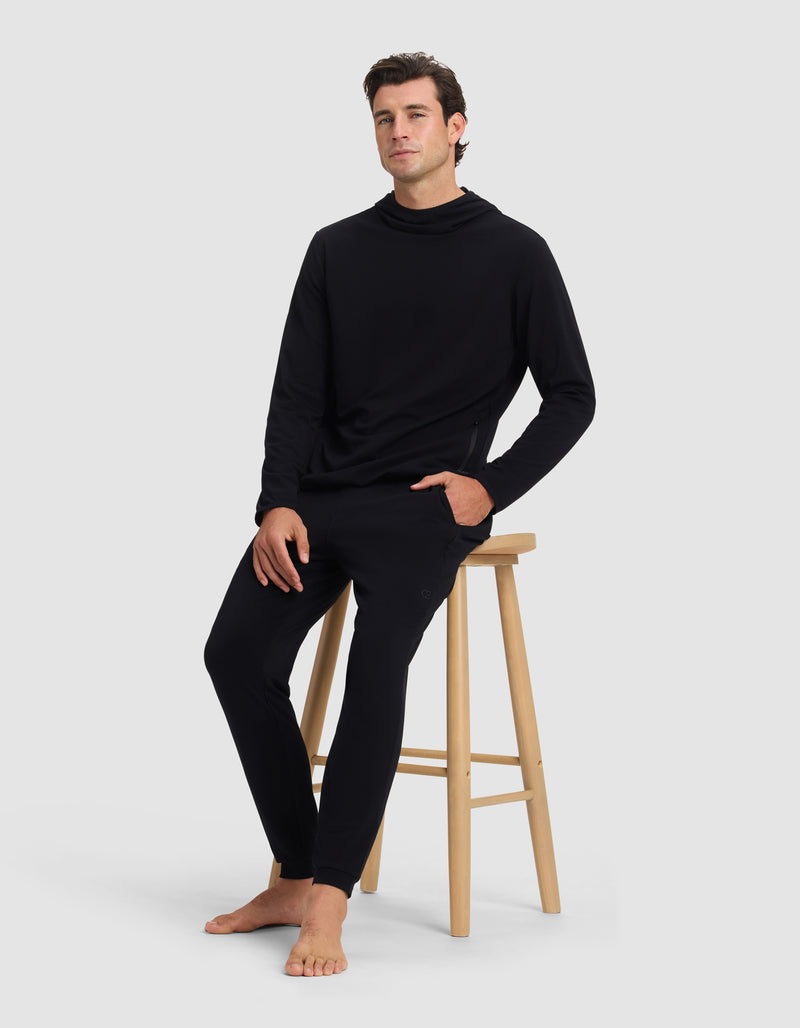 A man wearing the Cozy Earth Men's Brushed Bamboo Recovery Hoodie sits barefoot in black jogger pants on a wooden stool against a plain light gray background.