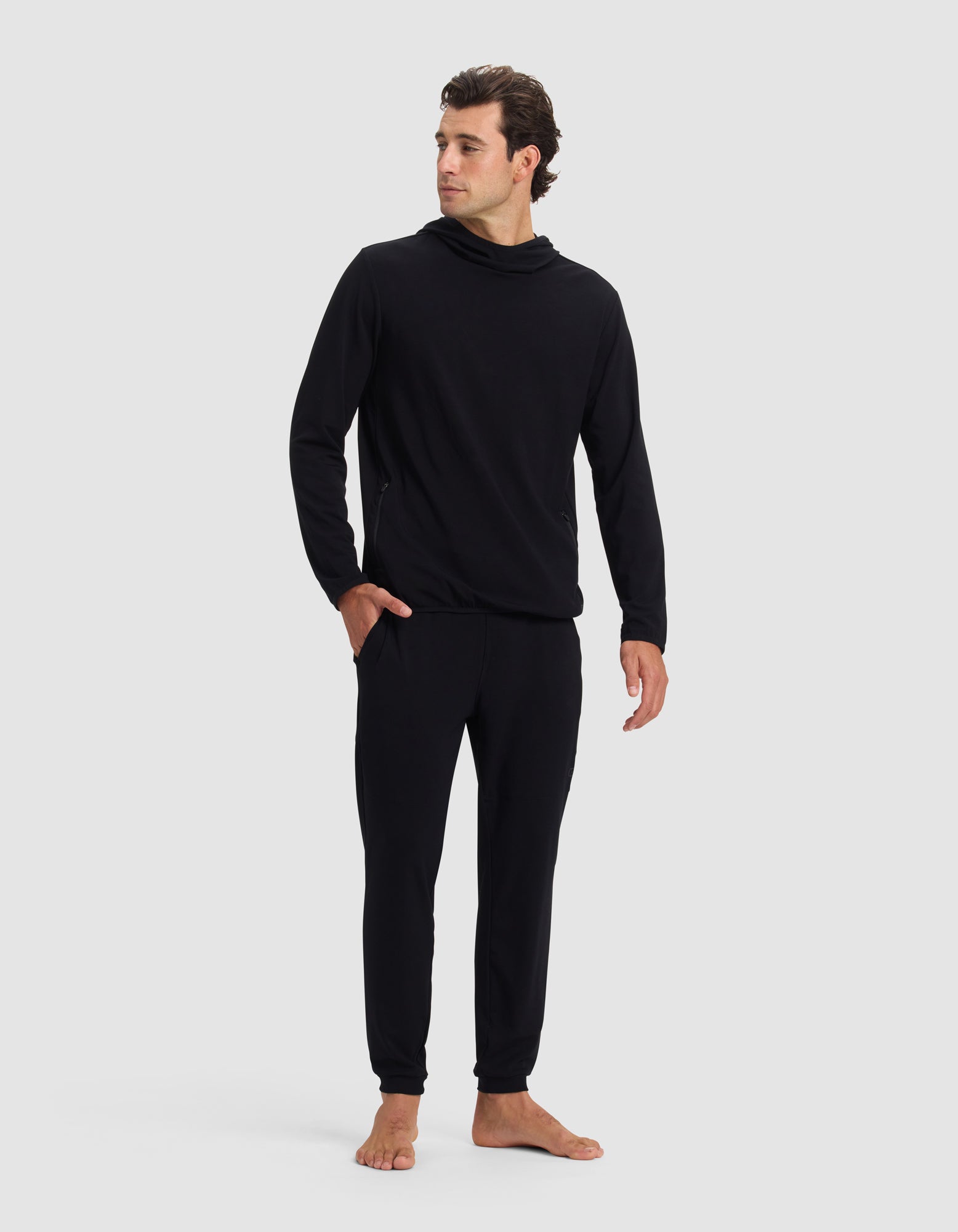A man stands barefoot against a plain background, wearing the Cozy Earth Men's Brushed Bamboo Recovery Hoodie & Jogger Set in black, featuring a loose-neck hoodie and jogger pants. He looks slightly to the side with a relaxed posture.