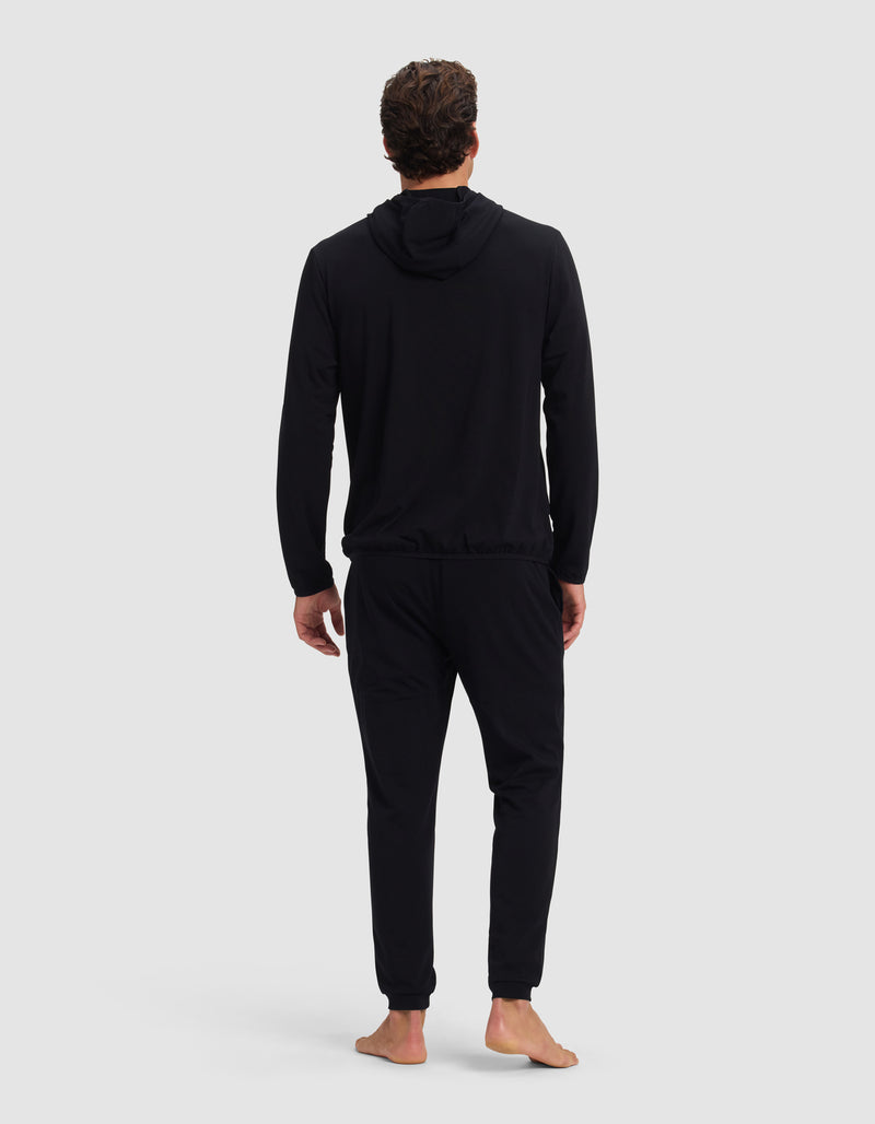 A man stands barefoot facing away, wearing the Cozy Earth Men's Brushed Bamboo Recovery Hoodie & Jogger Set in black, against a plain light gray background.