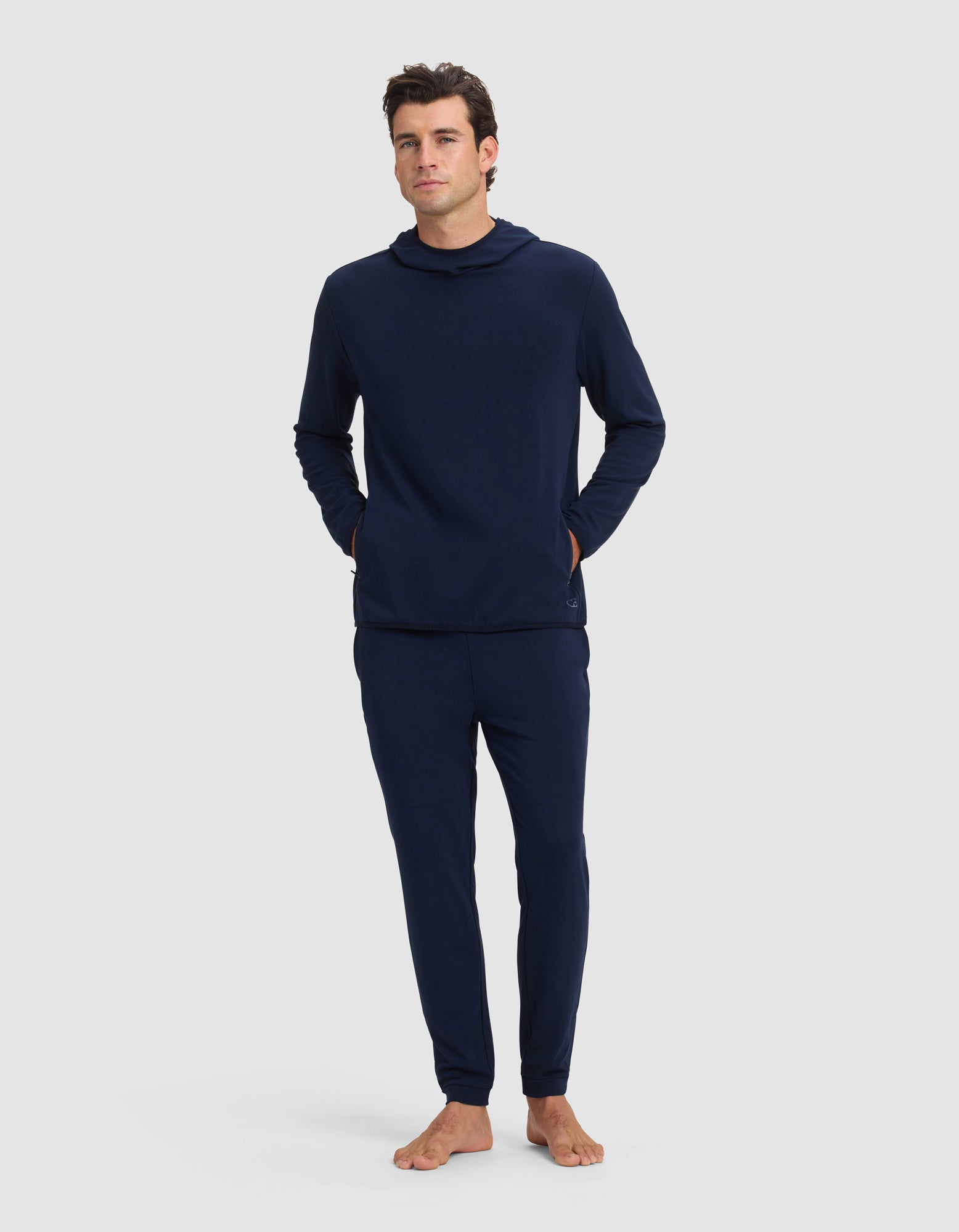 A man stands barefoot against a light gray background, wearing the Cozy Earth Men's Brushed Bamboo Recovery Hoodie & Jogger Set in dark blue, with his hands in his pockets.