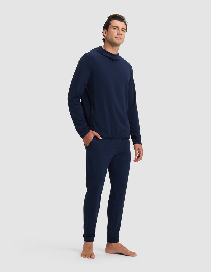 A man with short brown hair stands barefoot against a plain background, wearing the Cozy Earth Men's Brushed Bamboo Recovery Hoodie & Jogger Set in dark blue. He gazes slightly upward with a neutral expression.