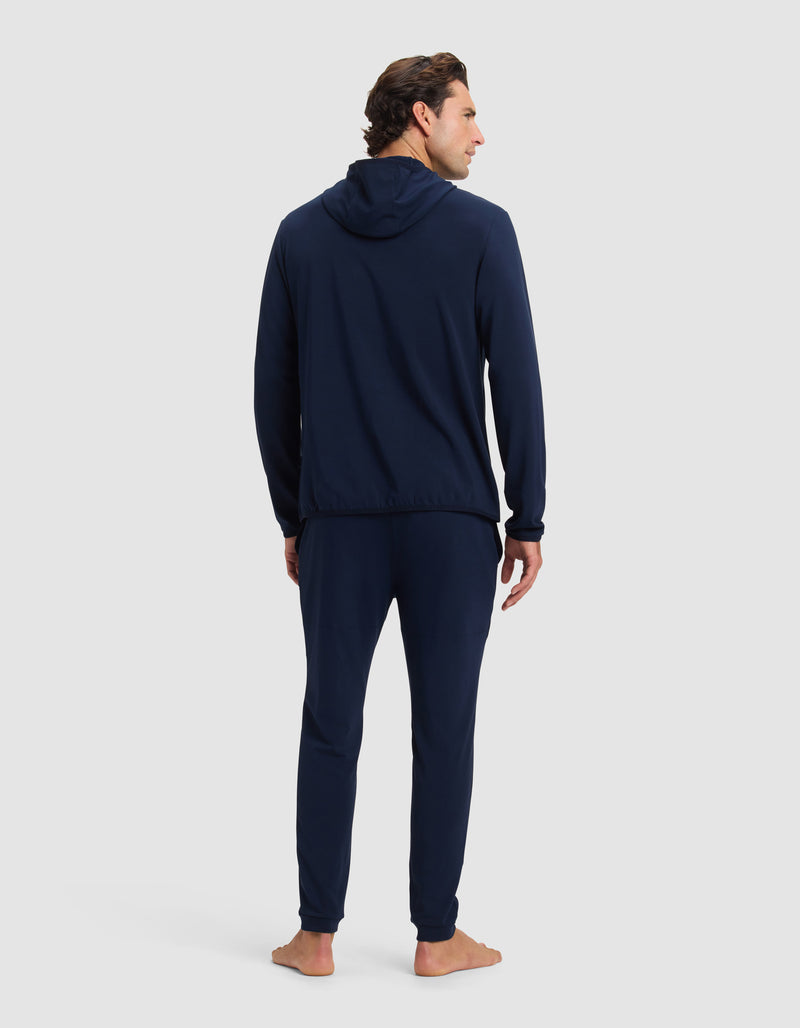 A man stands barefoot facing away, wearing the Cozy Earth Men's Brushed Bamboo Recovery Hoodie & Jogger Set in navy blue, against a plain light gray background.