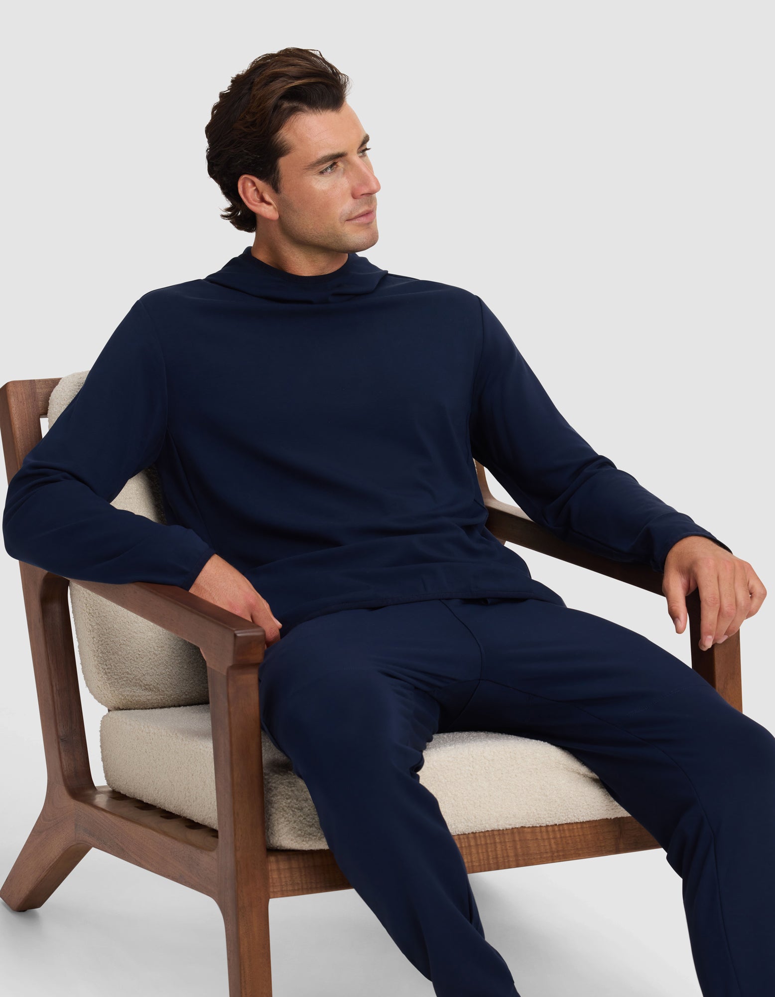 A man in a Cozy Earth Men's Brushed Bamboo Recovery Hoodie and matching pants sits on a cushioned wooden chair, looking left with a relaxed expression against a plain light background. |Color:Navy