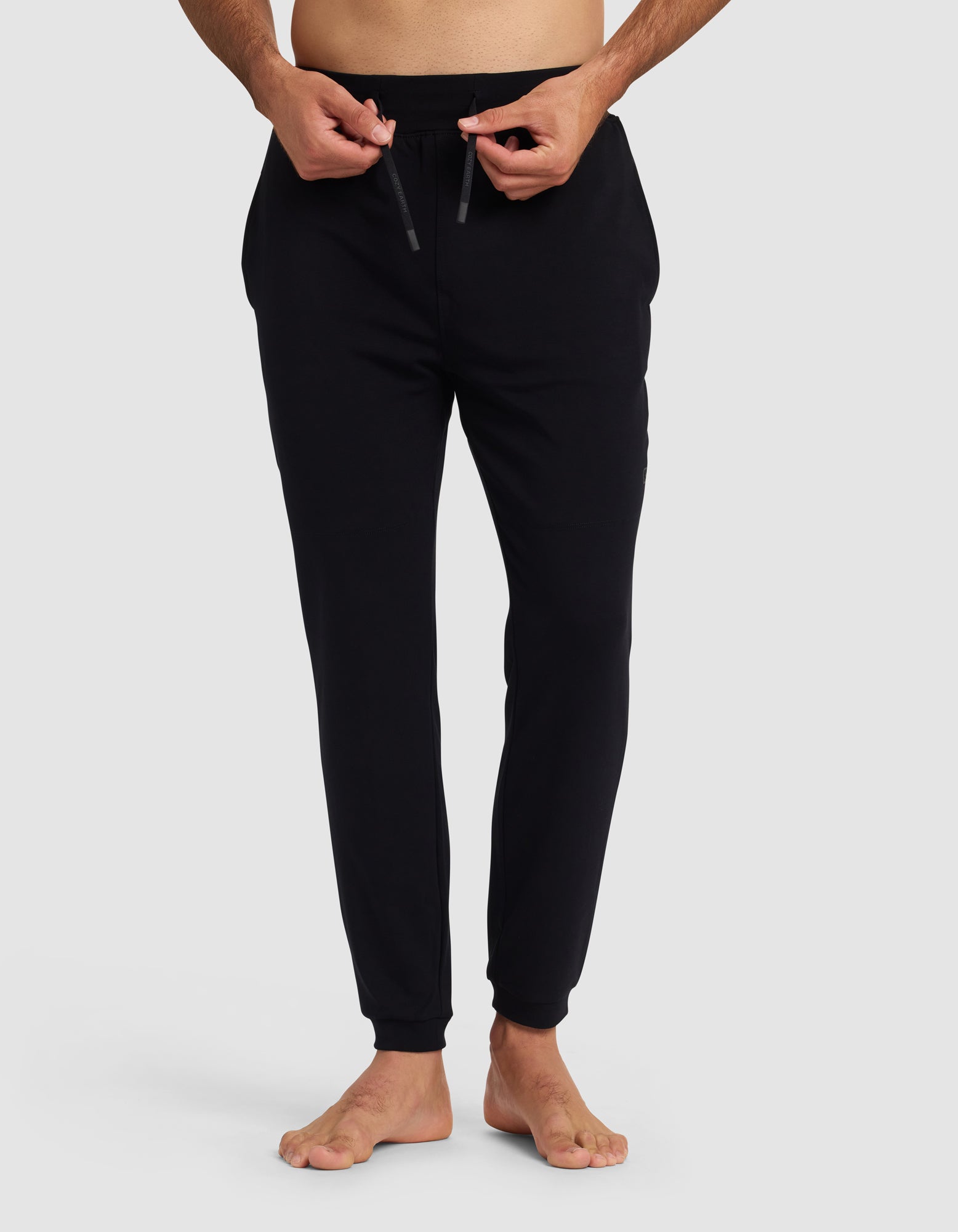 A person stands barefoot on a white background, wearing Cozy Earth's Men's Brushed Bamboo Recovery Jogger in black and adjusting the waistband drawstring. Their upper body is mostly out of frame.