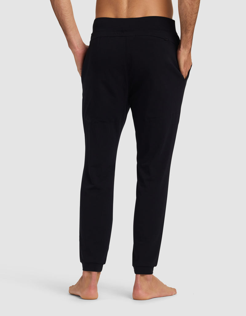 A barefoot man is shown from the back wearing Cozy Earth's Men's Brushed Bamboo Recovery Jogger, featuring a fitted waistband and cuffed ankles, with hands in pockets. The background is plain and light-colored.