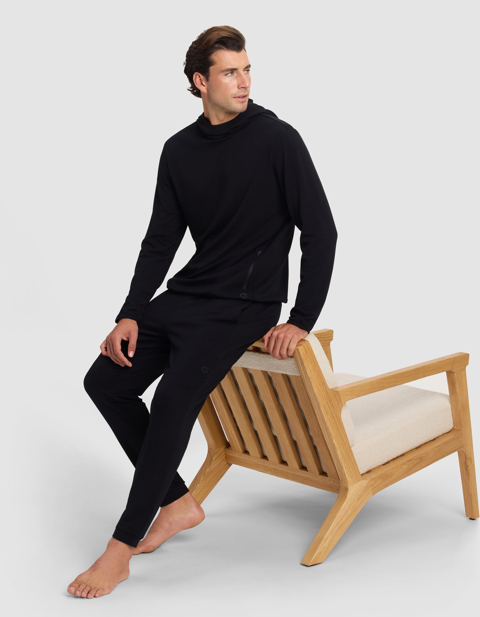 A man in Cozy Earth's Men's Brushed Bamboo Recovery Jogger, in black, sits barefoot on the arm of a modern wooden chair with cream cushions, set against a plain light gray background.