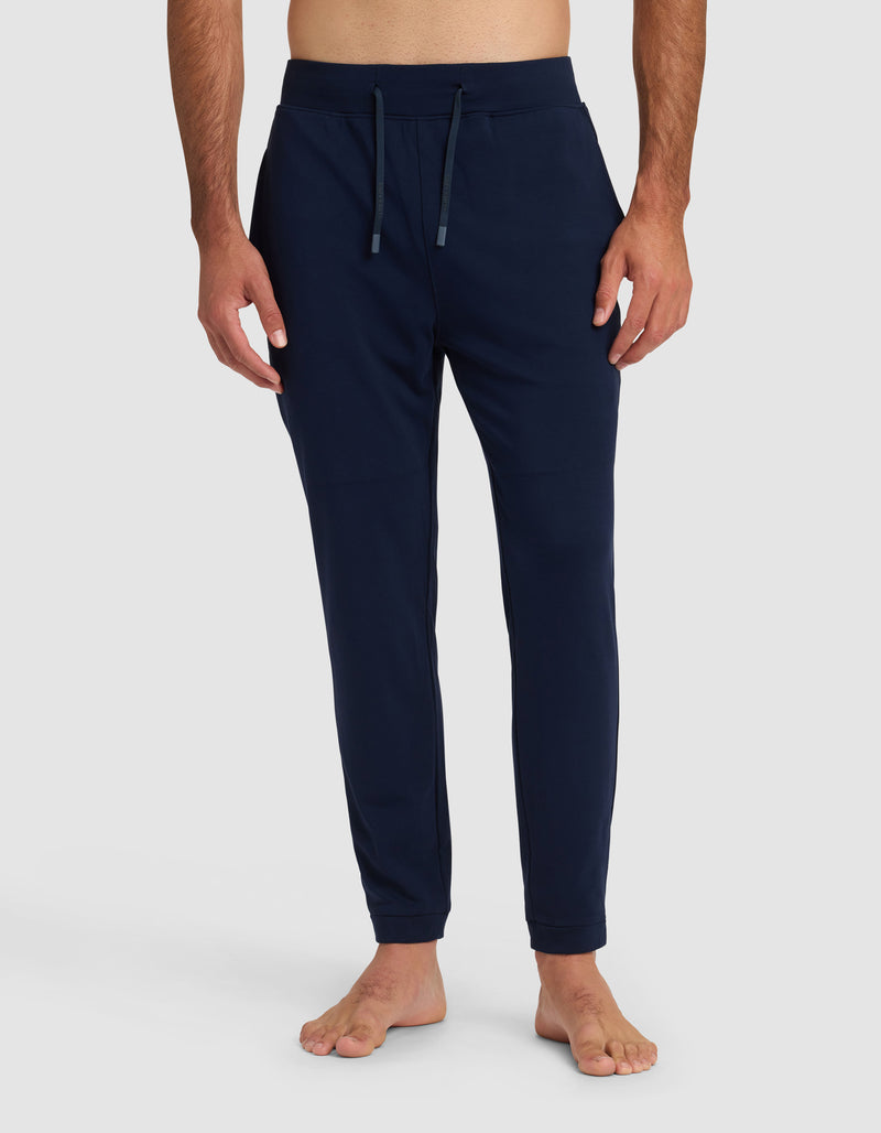 A person stands barefoot against a plain background, wearing Cozy Earth’s Men’s Brushed Bamboo Recovery Jogger in navy blue. The joggers feature a drawstring waistband, side pockets, and a slim fit with the torso not fully visible.