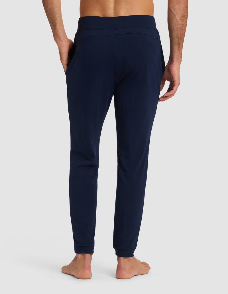 A barefoot man is shown from the back wearing Cozy Earth's Men's Brushed Bamboo Recovery Jogger in dark blue, with his hands in the side pockets, set against a plain light background.