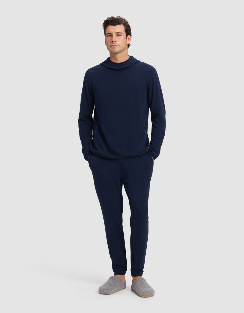 A man stands facing forward in a Cozy Earth Men's Brushed Bamboo Recovery Jogger set—navy blue hooded sweatshirt and jogger pants—with his hands in his pockets, wearing gray slippers, against a plain light background.