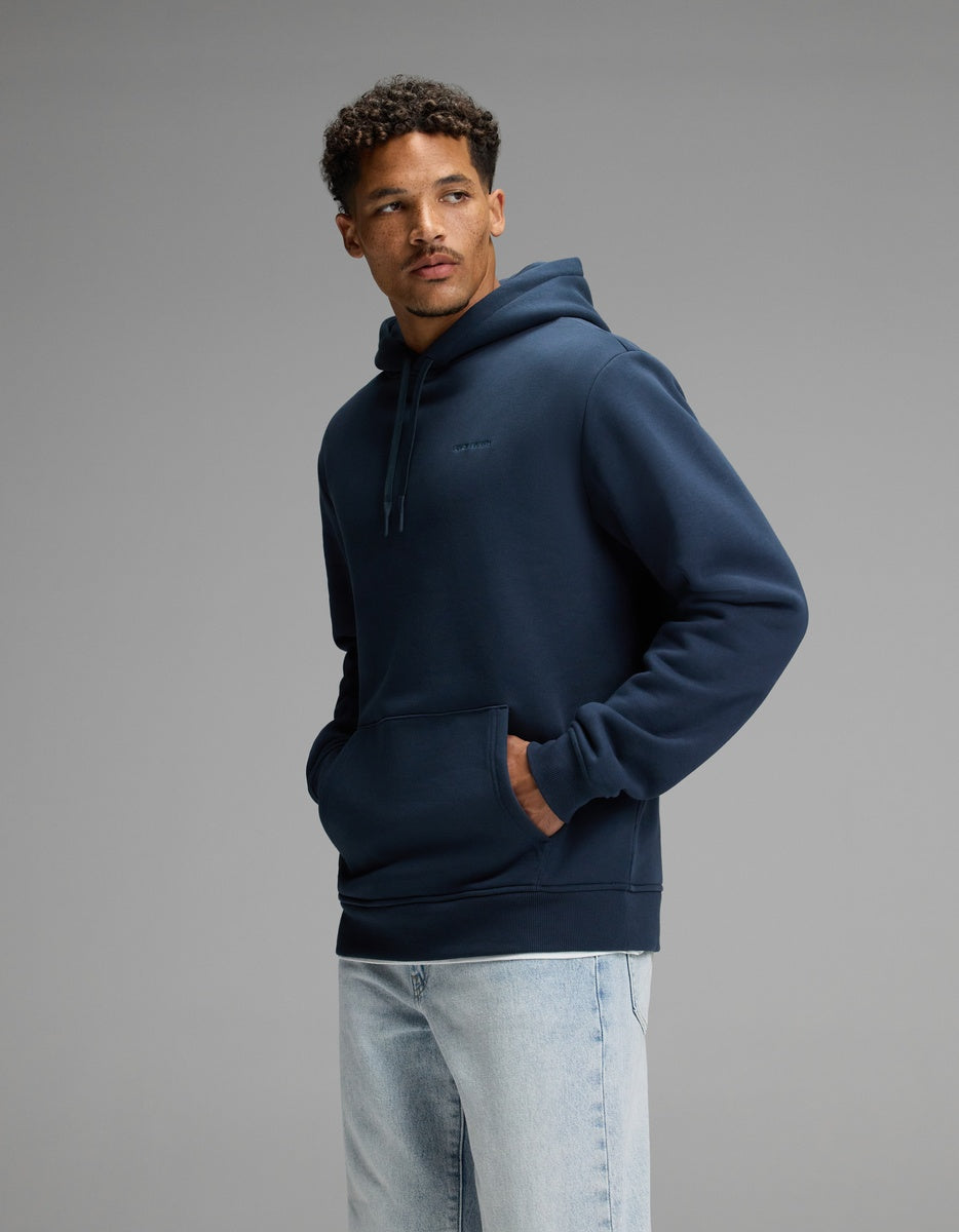 A man wearing the Cozy Earth Men’s Cityscape 2.0 Hoodie in Eclipse stands against a plain gray background with his hands in the front pocket, looking to the side, and styled with light blue jeans.|Color:Eclipse