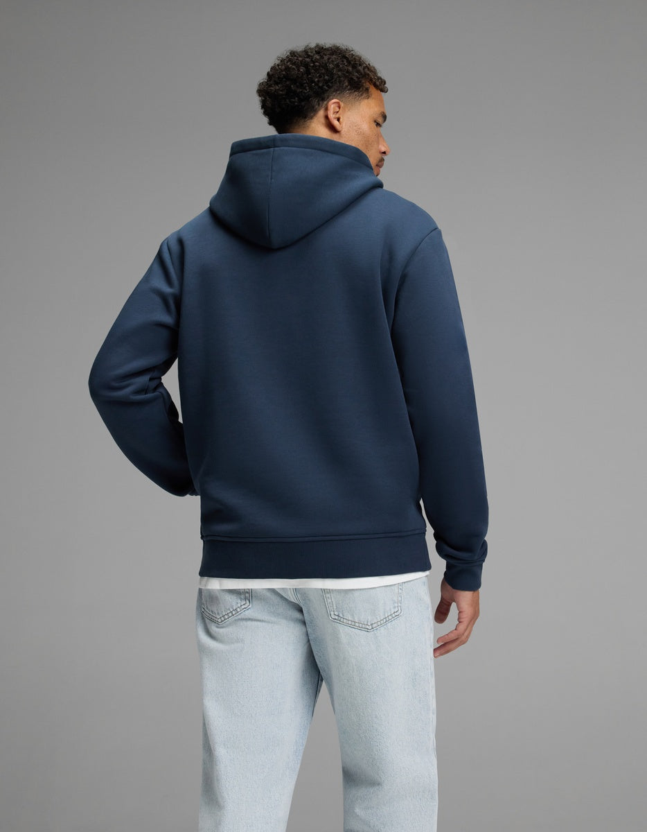 A person stands facing away, wearing the Cozy Earth Men’s Cityscape 2.0 Hoodie in navy blue and light blue jeans, against a plain gray background.