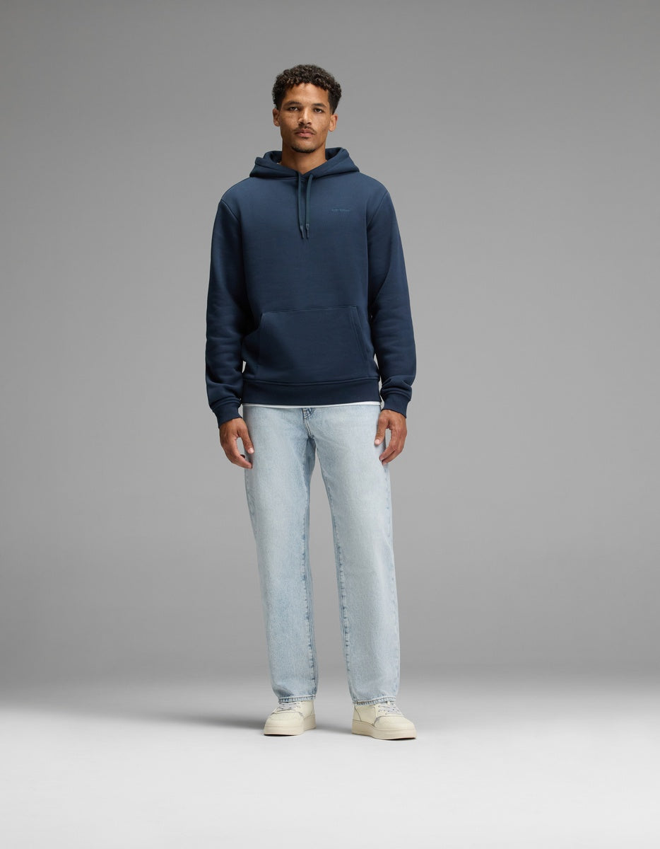 A man stands facing forward against a plain gray background, wearing the Cozy Earth Men’s Cityscape 2.0 Hoodie in navy blue, paired with light blue jeans and off-white sneakers.