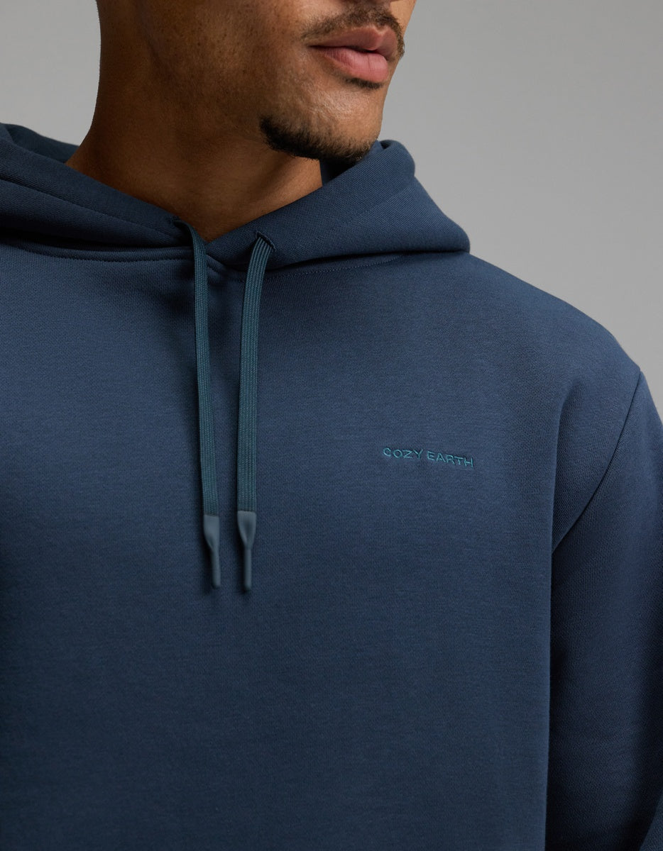 A person wearing the Men’s Cityscape 2.0 Hoodie by Cozy Earth in dark blue, shown from the shoulders to the mouth, with “Cozy Earth” embroidered on the chest and hood strings hanging down.