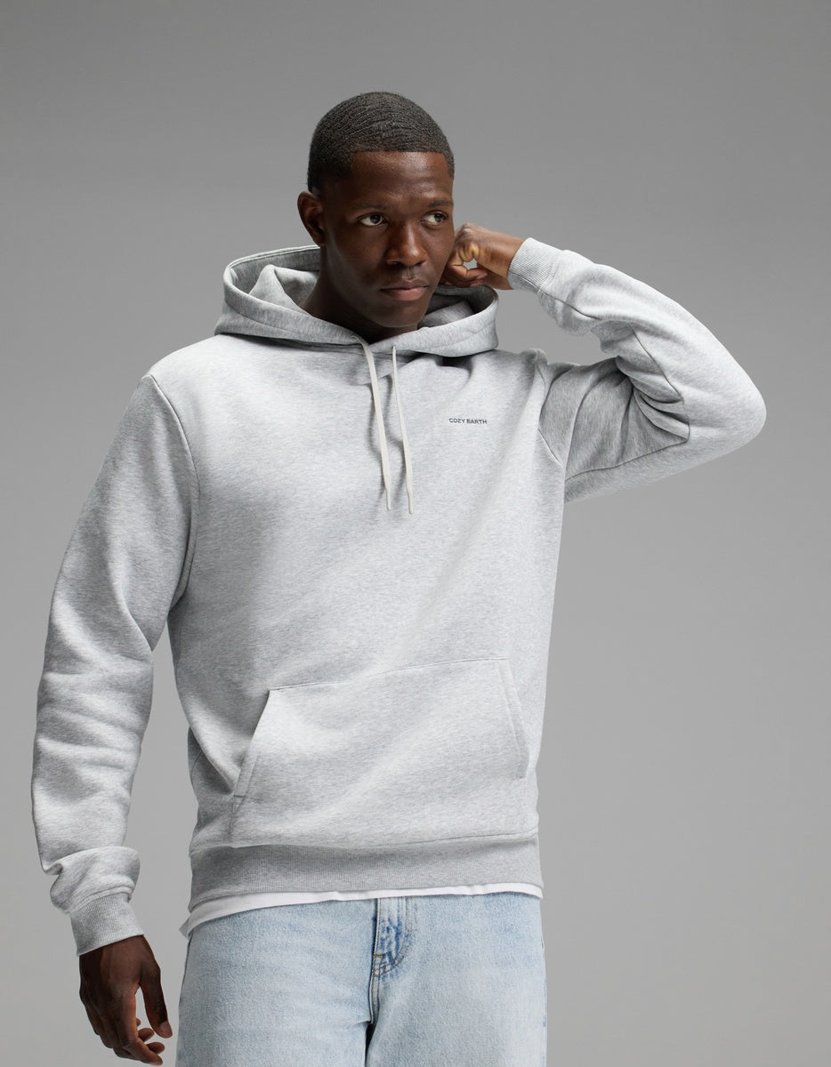 A man wearing the Cozy Earth Men’s Cityscape 2.0 Hoodie in Heather Grey and light blue jeans stands against a plain gray background, with one hand touching his head and the other relaxed by his side.