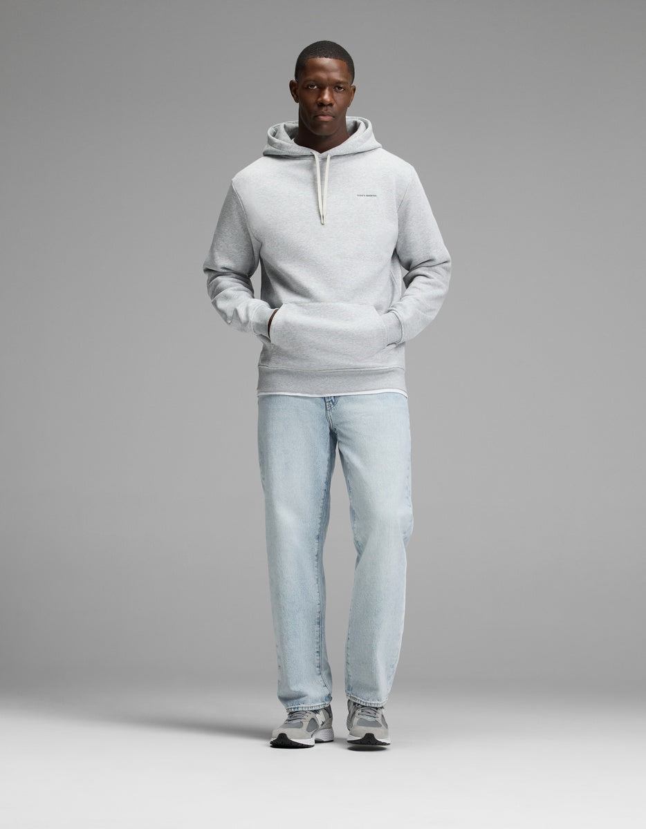 A man stands facing forward with his hands in the front pocket of a Cozy Earth Men’s Cityscape 2.0 Hoodie in light gray, paired with light blue jeans and gray sneakers against a plain gray background.
