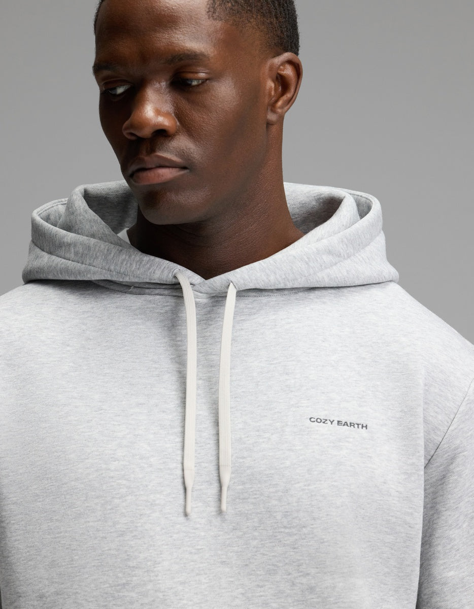 A man wearing the Cozy Earth Men’s Cityscape 2.0 Hoodie in light gray, featuring white drawstrings and “COZY EARTH” text on the chest, looks down with a neutral expression against a plain gray background.
