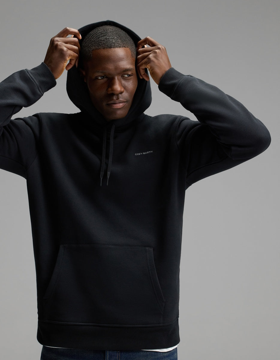 A man wears the Cozy Earth Men’s Cityscape 2.0 Hoodie in Jet Black, lifting the hood over his head as he stands against a plain gray background, gazing to the side with a neutral expression.