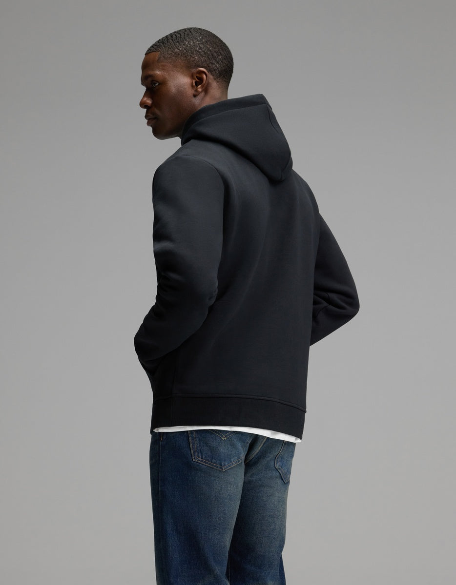 A person wearing the Cozy Earth Men’s Cityscape 2.0 Hoodie in black and blue jeans stands with hands in pockets, facing away and glancing to the side against a plain gray background.