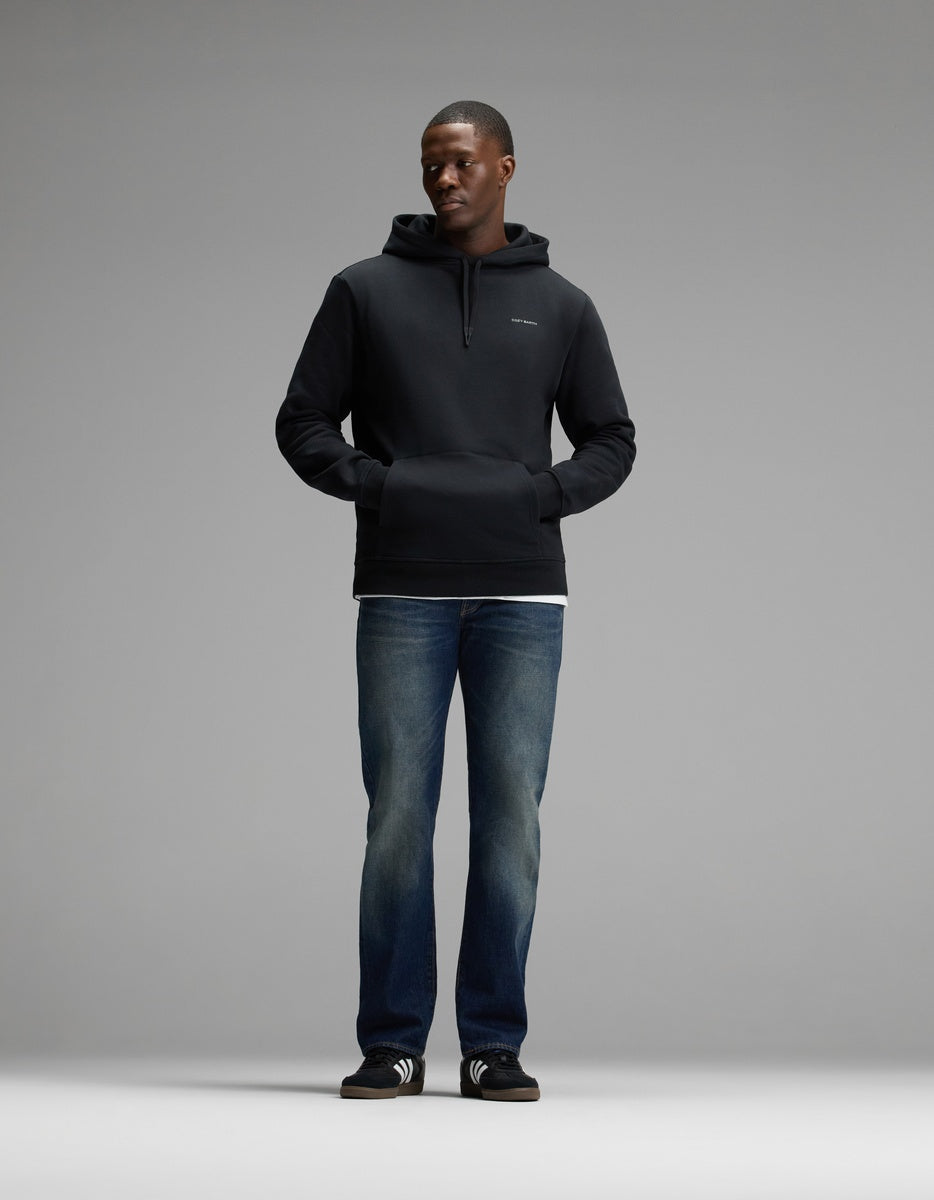 A man stands facing forward, wearing the Cozy Earth Men’s Cityscape 2.0 Hoodie in black, paired with blue jeans, black slide sandals with white stripes, and a white shirt visible at the hem. The background is plain and gray.