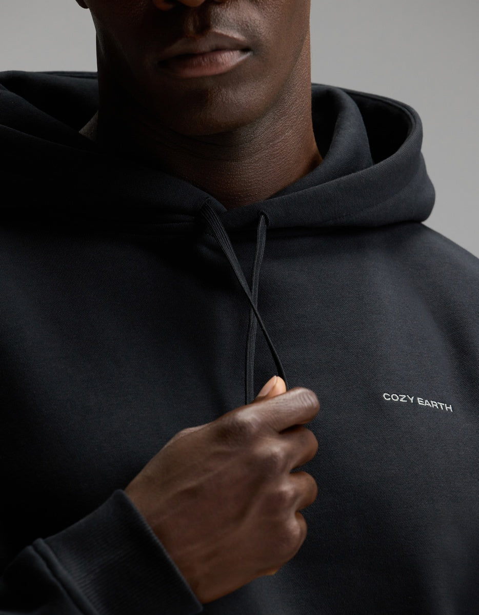 A person wearing the Cozy Earth Men’s Cityscape 2.0 Hoodie in black, featuring the “COZY EARTH” logo, holds one of the drawstrings. The photo is cropped to show only the lower half of their face and upper torso.