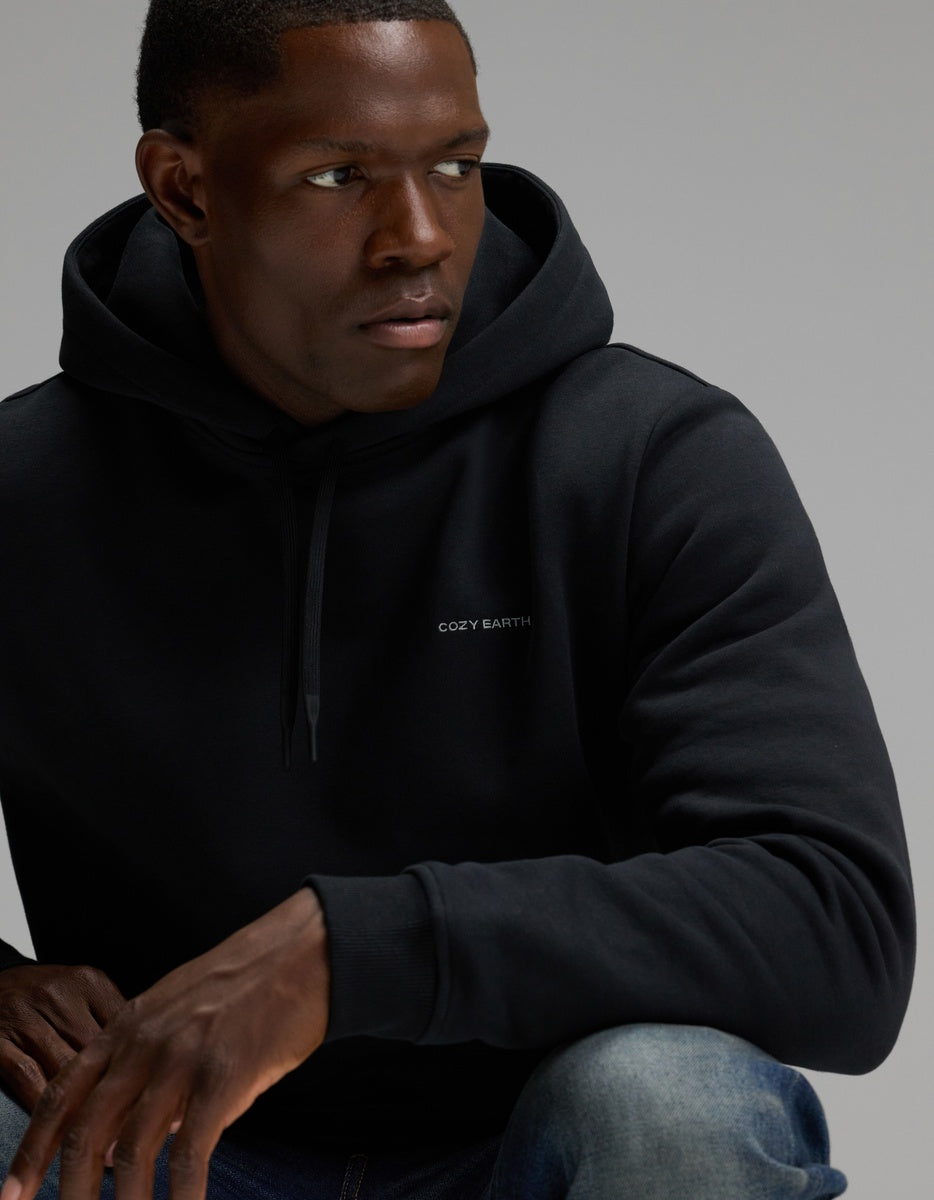 A man wearing the Men’s Cityscape 2.0 Hoodie by Cozy Earth, featuring the brand’s text on the front, looks to his left with one arm resting on his knee against a plain background.