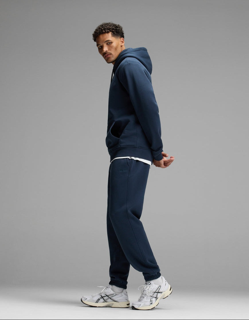 A man models the Cozy Earth Men’s Cityscape 2.0 Hoodie & Sweatpant Set in navy, paired with a white T-shirt and white sneakers, standing sideways against a plain gray background with his hands behind his back and looking over his shoulder.