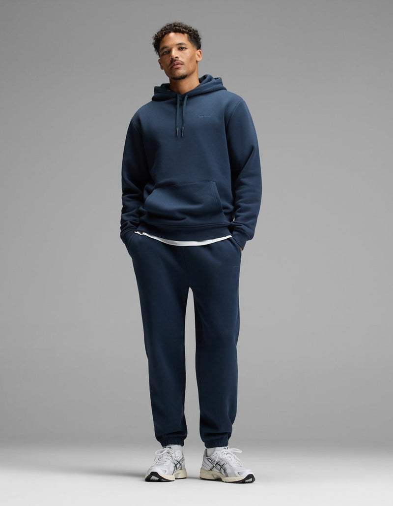 A person stands against a plain gray background, wearing the Cozy Earth Men’s Cityscape Hoodie & Sweatpant Set in Eclipse, paired with white sneakers and a white undershirt. Their hands are in their pockets and they are looking straight ahead.