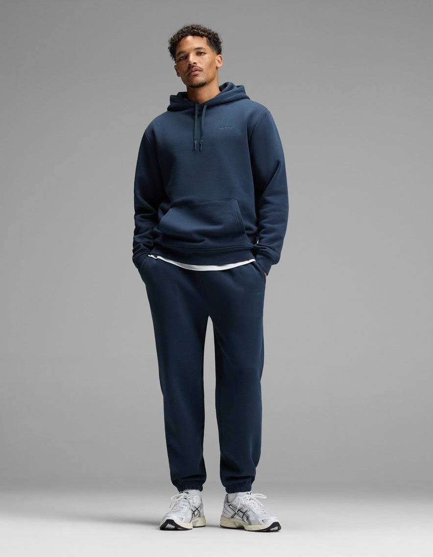 A person stands against a plain gray background, wearing the Cozy Earth Men’s Cityscape 2.0 Hoodie & Sweatpant Set in Eclipse, paired with white sneakers and a white undershirt. Their hands are in their pockets and they are looking straight ahead.|Color:Eclipse