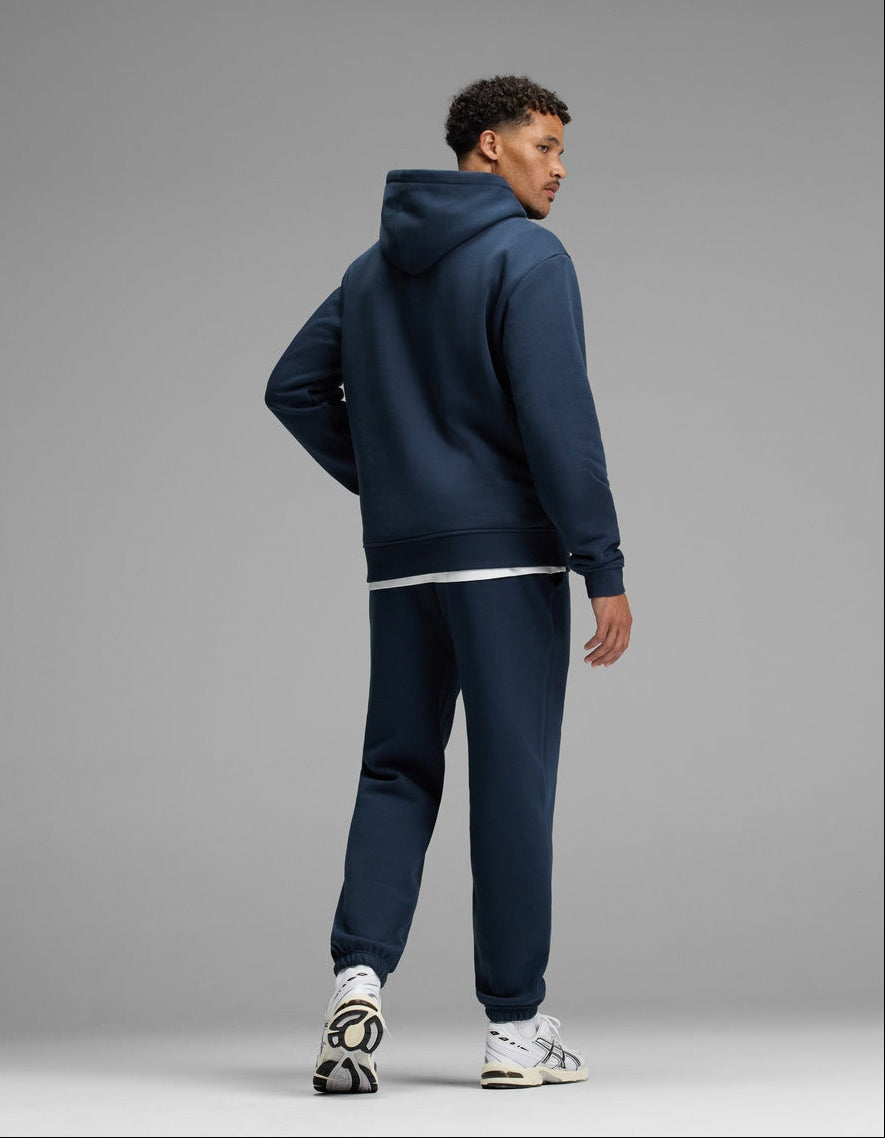A man stands facing slightly away, wearing the Cozy Earth Men’s Cityscape 2.0 Hoodie & Sweatpant Set in navy blue with white sneakers against a plain gray background.