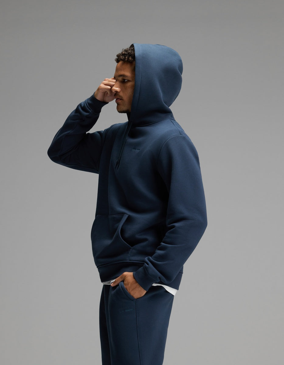 A person dressed in the Cozy Earth Men’s Cityscape 2.0 Hoodie & Sweatpant Set in navy stands in profile against a plain gray background, with one hand touching their forehead.