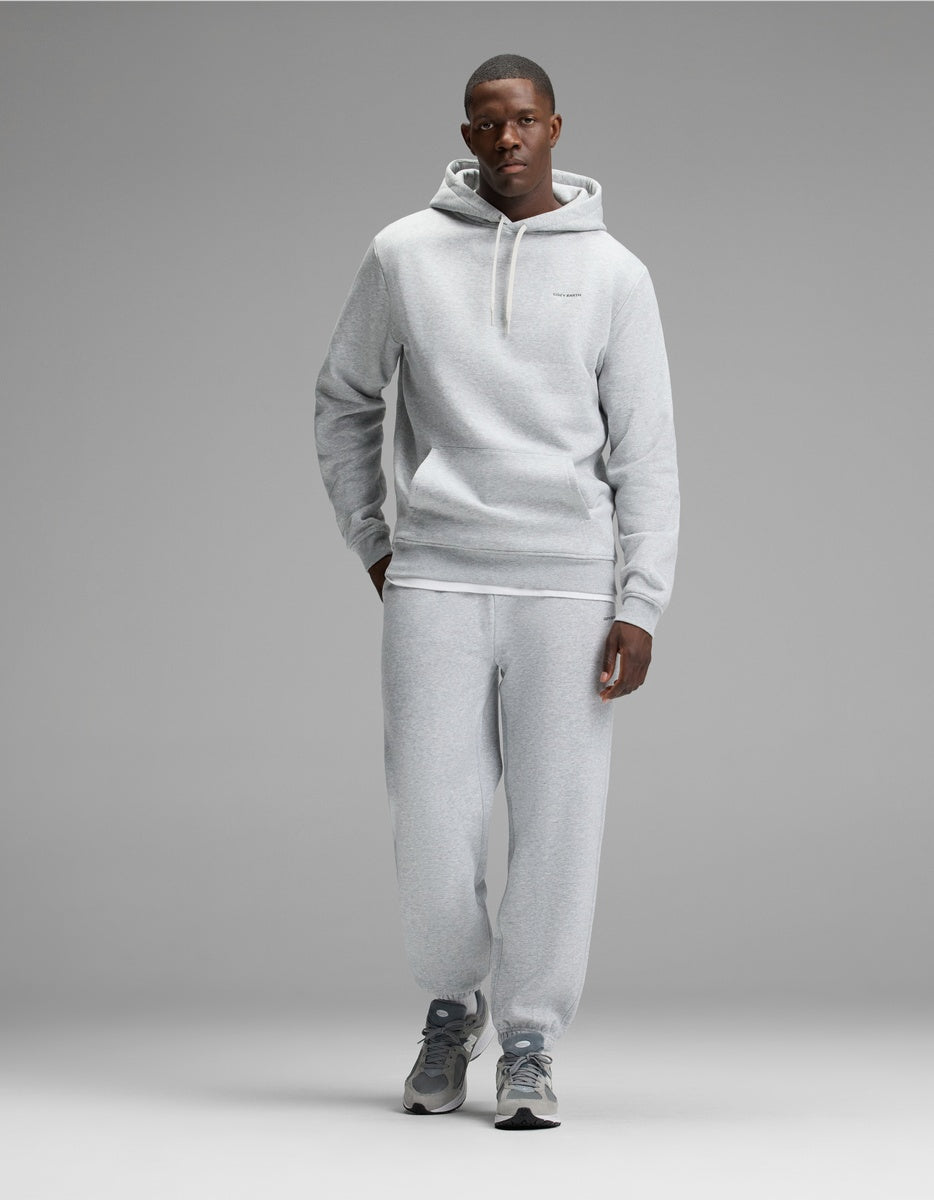 A man stands against a gray backdrop wearing the Cozy Earth Men’s Cityscape 2.0 Hoodie & Sweatpant Set in Heather Grey, paired with gray athletic shoes. His left hand is in his pocket, and he faces forward with a neutral expression.
