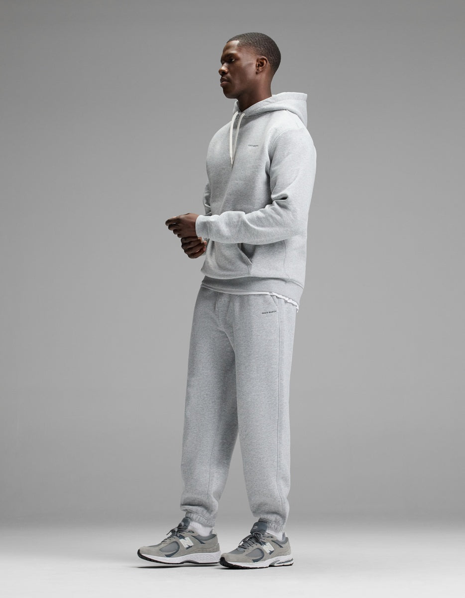 A person stands in profile against a gray background, wearing the Cozy Earth Men’s Cityscape 2.0 Hoodie & Sweatpant Set paired with gray sneakers. The outfit looks casual and comfortable.