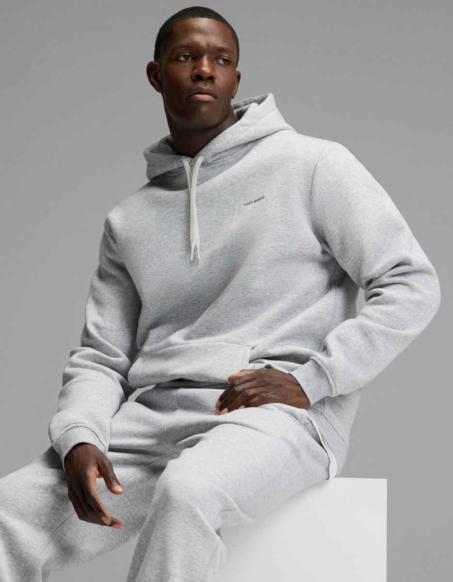 A man wearing the Cozy Earth Men’s Cityscape 2.0 Hoodie & Sweatpant Set in light gray sits on a white block against a plain gray background, looking to the side with a neutral expression.