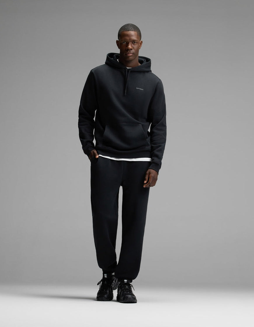 A man wearing Cozy Earth’s Men’s Cityscape 2.0 Hoodie & Sweatpant Set in Jet Black stands against a plain gray background, facing the camera with a neutral expression.
