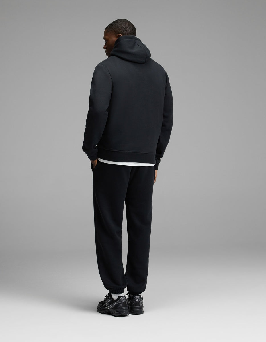 A person stands facing away, wearing the Cozy Earth Men’s Cityscape 2.0 Hoodie & Sweatpant Set in black against a minimal, plain gray background.