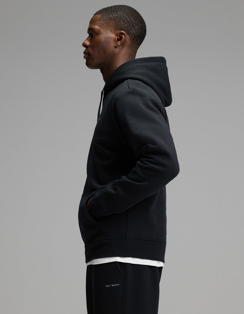 A man wearing the Cozy Earth Men’s Cityscape 2.0 Hoodie & Sweatpant Set stands in profile against a plain gray background, his hands in the hoodie pocket as he looks forward.
