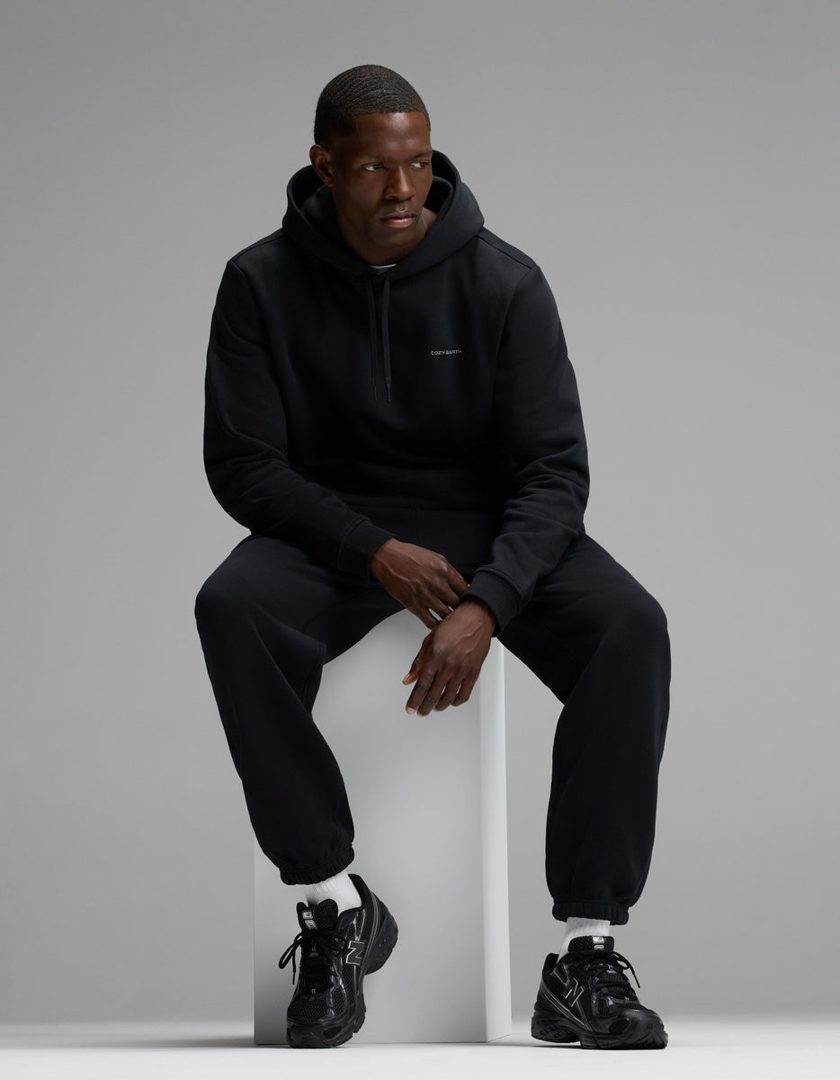 A man wearing the Cozy Earth Men’s Cityscape 2.0 Hoodie & Sweatpant Set sits on a white block against a gray background, paired with black sneakers and white socks.