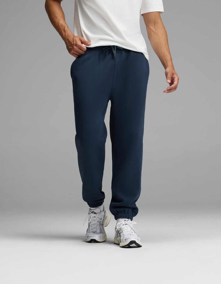 A person wearing a white T-shirt, white sneakers, and the Cozy Earth Men’s Cityscape 2.0 Sweatpant in navy blue stands against a plain gray background. The image is cropped to show the individual from the shoulders down.|Color:Eclipse