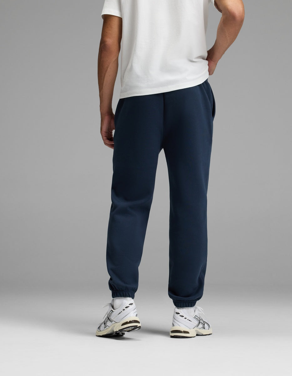 A person stands facing away, wearing a white T-shirt, Cozy Earth’s Men’s Cityscape 2.0 Sweatpant in navy blue, and white sneakers. The background is plain and gray.
