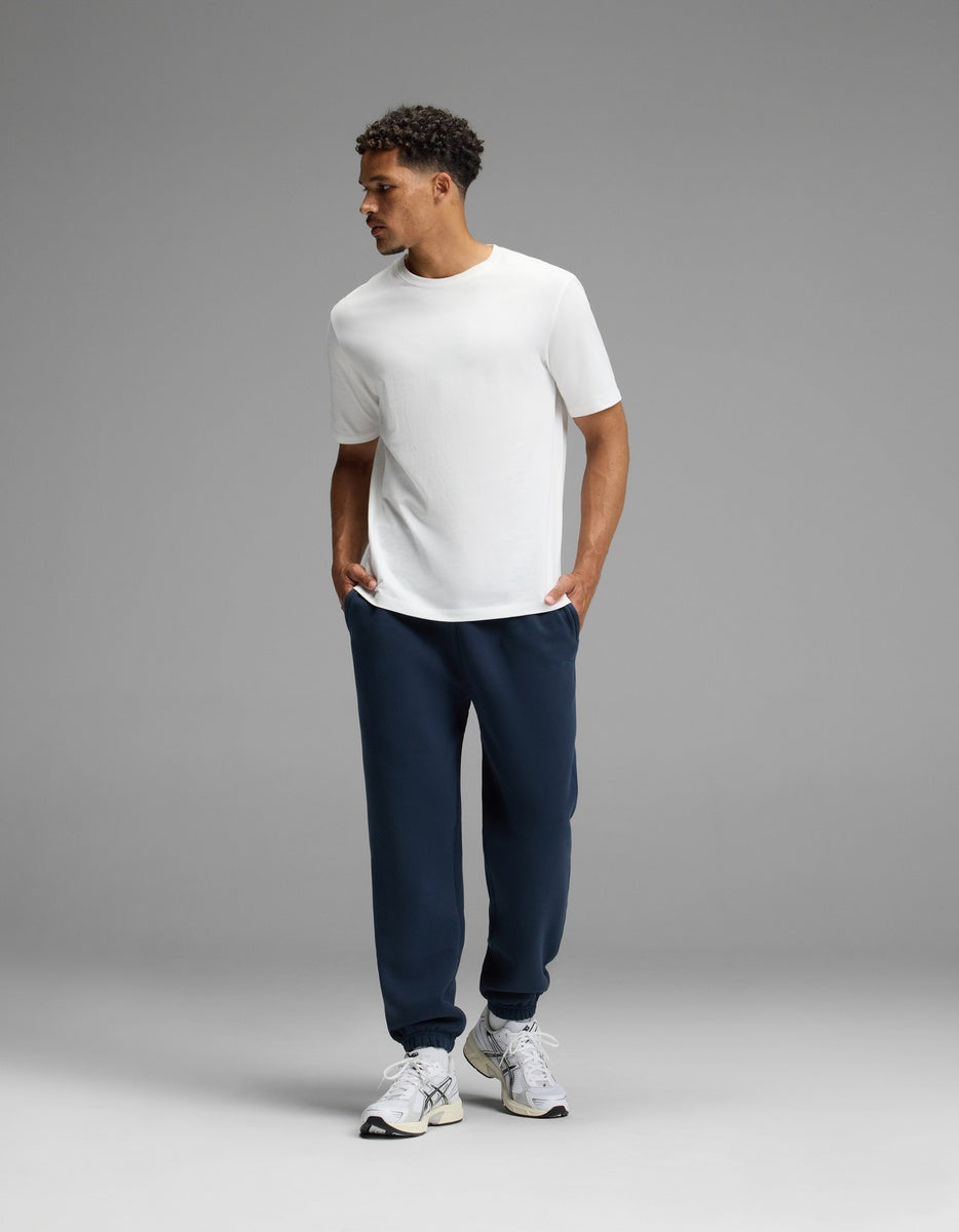 A man stands against a gray background, wearing a white t-shirt, white sneakers, and Cozy Earth’s Men’s Cityscape 2.0 Sweatpant in navy blue, with his hands in his pockets and looking to the side.