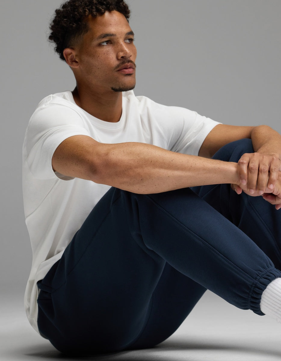 A man with short curly hair sits on the floor, arms resting on his knees, wearing a white t-shirt and Cozy Earth’s Men’s Cityscape 2.0 Sweatpant in navy. He looks off to the side with a neutral expression against a plain gray background.