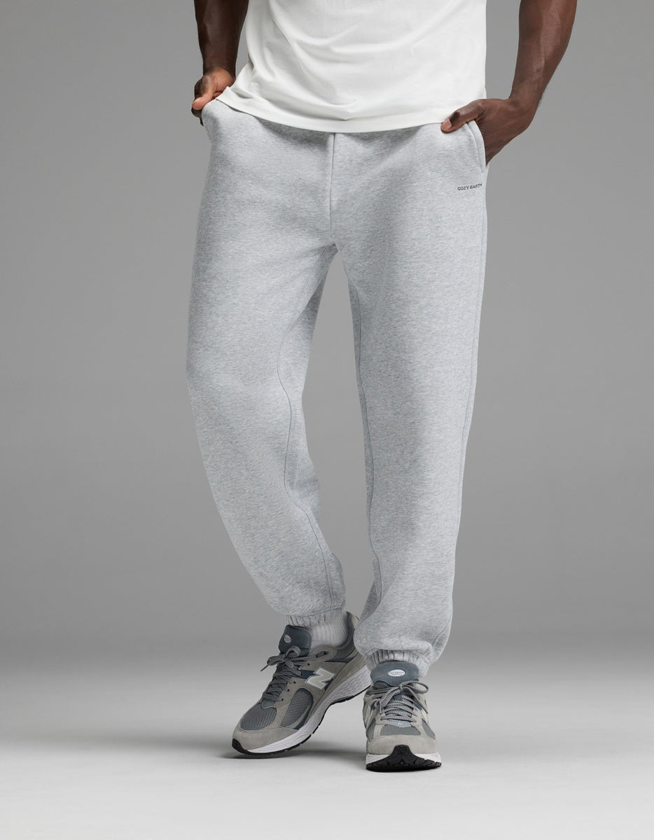 A person wearing a white shirt, Cozy Earth Men’s Cityscape 2.0 Sweatpant in light gray, and gray sneakers stands with hands in pockets against a plain gray background.