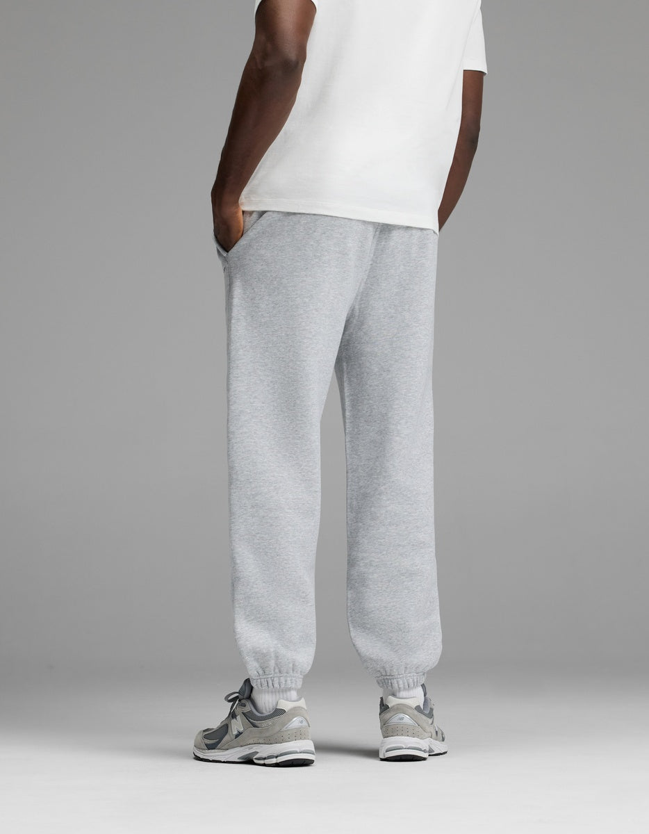A person stands with hands in pockets, wearing a white T-shirt, Cozy Earth Men’s Cityscape 2.0 Sweatpant in light gray, and gray sneakers. The background is plain gray. The image is shown from behind the knees up.